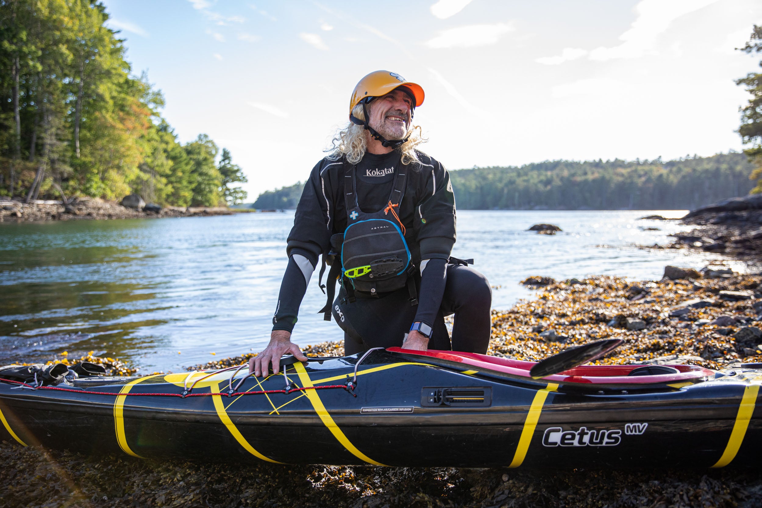 Person with a kayak and PFD mounted knife on a rocky shoreline with trees in the background